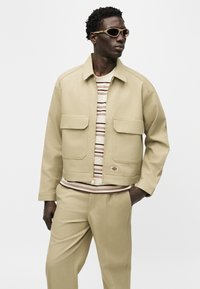 Άνδρας που φοράει μπεζ σακάκι και παντελόνι με ριγέ πουκάμισο, τα χέρια στις τσέπες, κοιτάζοντας δεξιά, σε απλό λευκό φόντο.