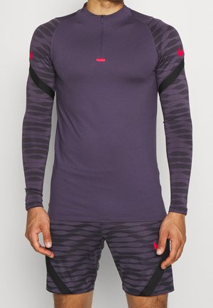 Hombre con camiseta deportiva de manga larga color morado oscuro con mangas estampadas en negro y pantalones cortos a juego con logotipos rojos de Nike.