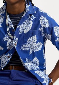 Niebieska koszula z krótkim rękawem z białym tropikalnym wzorem liści, zapinana na guziki z przodu, z kieszenią na piersi. Nosi się ją na pasiastą granatową koszulk�ę.