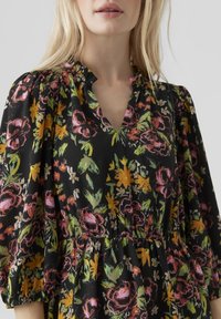 Černé květinové šaty s výstřihem do V, zdobené živými růžovými, oranžovými a zelenými květy. Materiál je měkký, s nařaseným pasem a nabíranými rukávy.