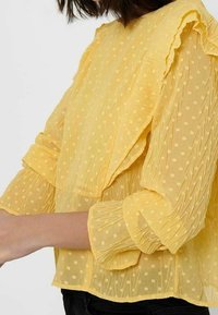 Gelbe Bluse mit Pünktchen aus durchsichtigem Stoff. Verfügt über Rüschendetails, lange Ärmel und strukturierte Streifen, mit einem lockeren Schnitt.