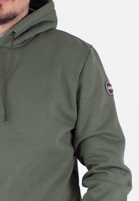 Felpa verde oliva con tasca a canguro, cappuccio con cordino e patch rotondo sulla manica sinistra. Tessuto morbido e testurizzato.