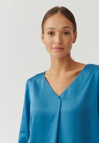Blusa di raso blu con scollatura a V, maniche lunghe e una consistenza morbida e liscia. Presenta un delicato pizzico al centro anteriore.