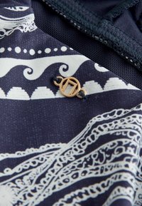Tissu bleu marine avec des motifs paisley et ondulés blancs complexes, avec une petite boucle circulaire en or attachée avec un fil noir.