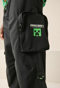 Fekete cargo nadrág zippzáras zsebbel, zöld Minecraft Creeper logóval és kontrasztos varrással, könnyű szövetből készült.
