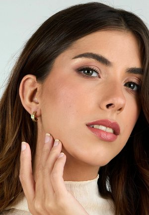 Orecchini a cerchio in oro con design attorcigliato e piccole accentuazioni quadrate, abbinati a un top beige morbido e texturizzato. La pelle appare liscia con un trucco naturale.