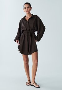 Rjava ohlapna obleka s srajco, z dolgimi rokavi, gumbi spredaj in pasom z vrvico. Kombinirano s črnimi ravnimi sandali.