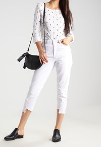 Weiße, cropped Jeans mit fransigem Saum, graues Langarmshirt mit kleinem schwarzen Herzmuster, schwarze Leder-Mules und eine schwarze Umhängetasche.