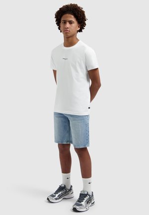 Jonge man staat met handen achter zijn rug, draagt een wit T-shirt, lichte denim shorts, witte sokken en grijze sportschoenen op een witte achtergrond.