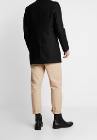 Blazer noir sur mesure aux épaules structurées, associé à un pantalon crème à revers et des bottines en cuir noir.