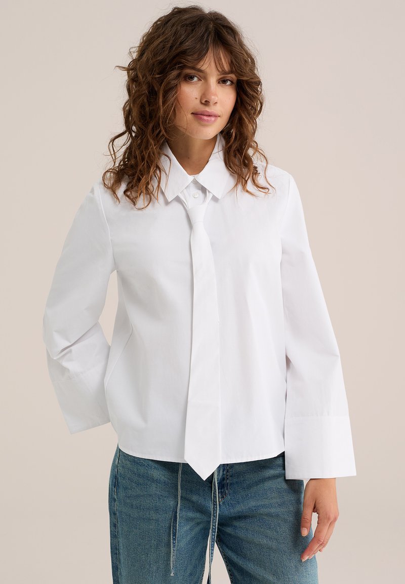 Femme aux cheveux bruns bouclés portant une chemise blanche à manches longues boutonnée avec une cravate blanche assortie et un jean bleu sur un fond uni.