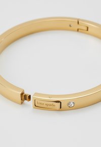 Bracelet en métal doré avec une surface lisse, arborant un petit accent en pierre claire et gravé "kate spade" sur le fermoir.