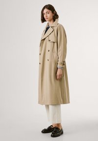 Beige trenchcoat med dubbelknäppta knappar, stor krage och sidofickor, kombinerat med vita byxor och svarta loafers.
