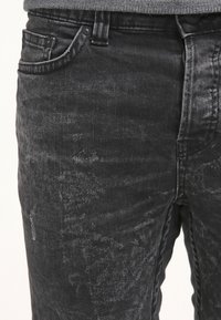 Jeans en denim noir avec une texture légèrement délavée, design à cinq poches et accents métalliques à l'avant. Les caractéristiques incluent une fermeture à bouton et des passants de ceinture.