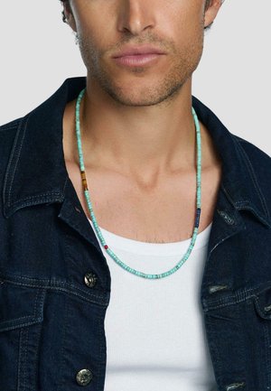 Mand iført en tyrkis perlekæde over en hvid tanktop og en mørk denimjakke, vist fra halsen til den øverste brystkasse.