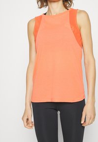 Vrouw in een mouwloze oranje sporttanktop en zwarte leggings, staande voor een effen witte achtergrond.
