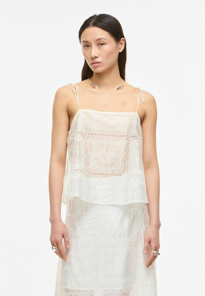 Jeune femme aux cheveux raides et foncés porte un haut blanc sans manches en dentelle et une jupe assortie, debout devant un fond blanc uni.