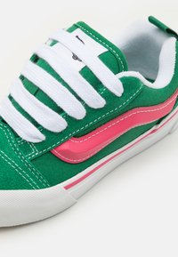 Grüner Stoff-Sneaker mit weißen Schnürsenkeln, pinken Lederakzenten und einer weißen Gummisohle. Verfügt über Kontrastnähte und ein minimales Design.