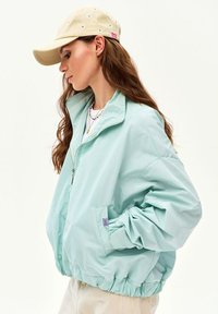 Woman with long hair wearing a light blue zip-up jacket, layered necklaces, beige pants, and a beige baseball cap with embroidered flowers.
