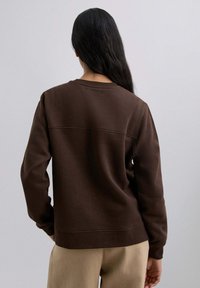 Sudadera marrón con un acabado texturizado, cuello redondo y mangas largas, con una costura en la parte de atrás. Combinada con pantalones de color claro.