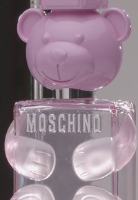 Flakon perfum Moschino z różowym korkiem w kształcie misia, przejrzysta szklana butelka oraz teksturowane różowe odciski łap po bokach, z wytłoczonym logo.