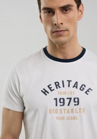 Balta trumpa rankovė t-shirt su tamsiai mėlynu apykakle. Ant jos atspausdintas tekstas tamsiai mėlyna ir auksine spalvomis: "PAVILDIKITE SAVO GYVENIMĄ. 1979 DIDELIS ŽVAIGŽDUTĖS UAB JŪSŲ DŽINSAI."