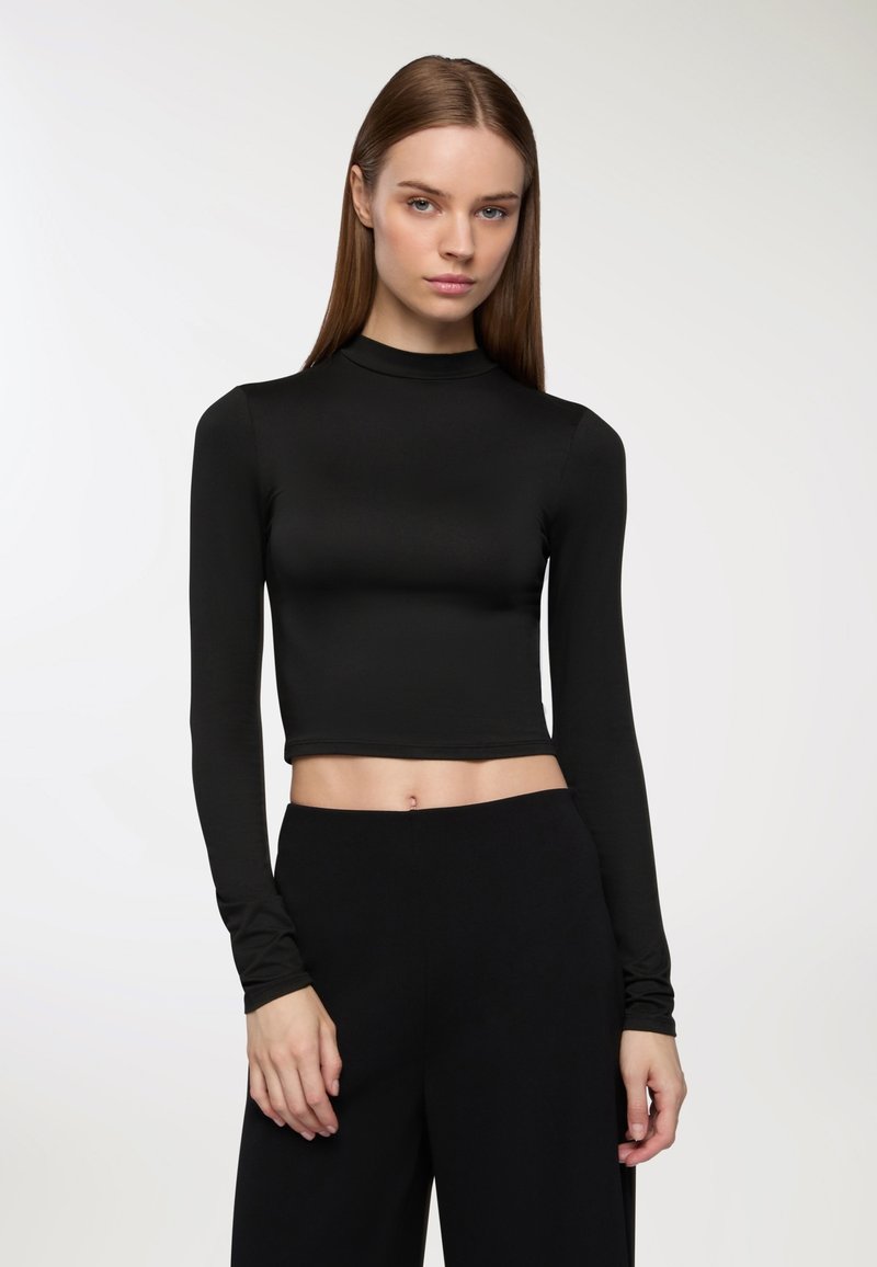 Crop top nero a maniche lunghe con collo alto. Tessuto liscio ed elastico. Abbinato a pantaloni neri a vita alta. Texture piatta; senza motivi o dettagli.