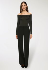 Body a maniche lunghe nera con spalle scoperte abbinato a pantaloni neri a vita alta con gamba larga e tacchi a punta neri. Texture lisce e aderenti.