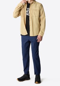 Camicia a maniche lunghe di colore tan, indossata sopra una maglietta nera, abbinata a pantaloni blu navy e scarpe da trekking nere con suole in gomma e lacci.