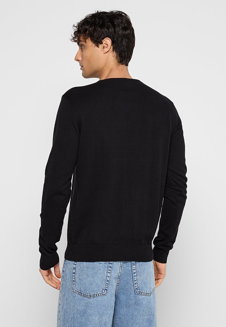 Schwarzer Strickpullover mit Rundhalsausschnitt, gerippten Bündchen und Saum; getragen über hellblauen Jeans. Minimalistisches Design mit strukturiertem Stoff.