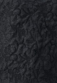 Tessuto nero con una superficie strutturata e un motivo arricciato, che presenta profondità variabili e una finitura opaca. Il materiale appare leggermente riflessivo alla luce.