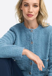 Veste texturée bleu clair avec un col rond, dotée de fermetures à boutons argentés et d'une poche frontale. Matière douce et duveteuse avec une coupe ajustée.