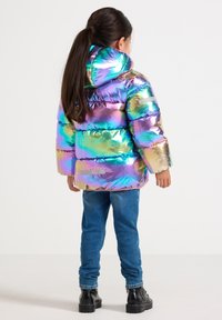 Enfant avec de longs cheveux noirs portant une doudoune iridescente brillante, un jean bleu et des bottes noires, se tenant de dos sur un fond uni.