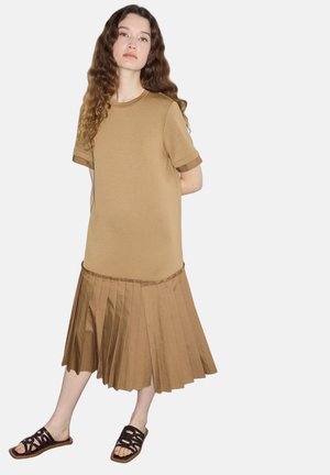 Junge Frau mit langen lockigen Haaren, die ein kurzärmeliges beiges Kleid mit plissiertem Rock und schwarzen offenen Sandalen trägt, steht vor einem weißen Hintergrund.