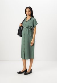 Abito premaman verde con bottoni, cintura in vita, maniche corte arrotolate e vestibilità comoda, abbinato a scarpe nere e una borsa.