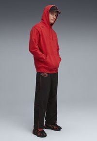 Rood capuchon sweatshirt met kangoeroezak, gecombineerd met zwarte broek met rode stiksels en logo. Draag met zwarte en rode sport schoenen.