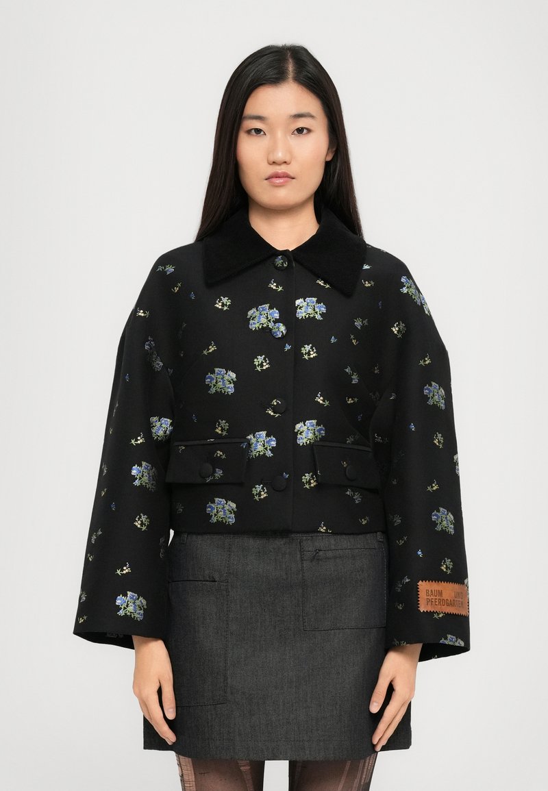 Μαύρο φλοράλ μπουφάν με μπλε και πράσινη κεντητική, μεγάλος γιακάς, μπροστινά κουμπιά και δύο πλαινές τσέπες. Συνδυάζεται με μια γκρίζα φούστα.