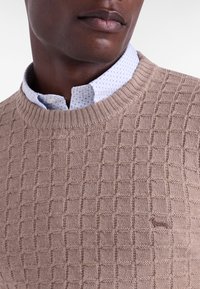 Beige knitted sweater with a textured square pattern. Features a ribbed neckline and a small embroidered logo on the chest.