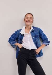 Teksas jakna klasičnega kroja, modre barve, z žepi spredaj; kombinirana z belo srajco z gumbi in črnimi oprijetimi kavbojkami.