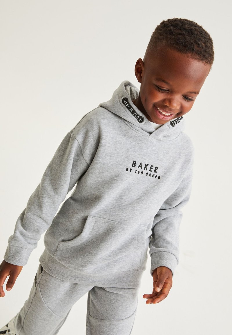 Chłopiec uśmiecha się i patrzy w dół, nosząc jasnoszarą bluzę z kapturem i dopasowane spodnie z tekstem "Baker by Ted Baker" na piersi.