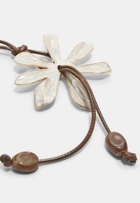 Ciondolo floreale realizzato in materiale chiaro e texture con dettagli dorati, collegato a un cordino intrecciato marrone con una perla decorativa.