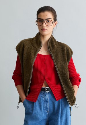 Junge Frau mit großen Brillen, die eine olivgrüne Fleeceweste, einen roten, gerippten Cardigan und hoch Taillen-Jeans in Blau trägt, mit den Händen in den Taschen.