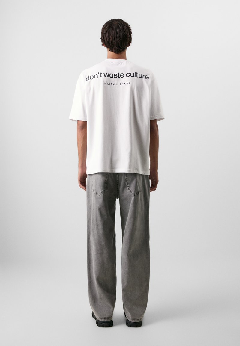 Hvid bomulds t-shirt med sort tekst "don’t waste culture" og "MAISON D'ART" på ryggen, parret med lysegrå bukser med vid pasform.