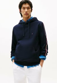 Granatowy hoodie z miękkiego materiału z przednią kieszenią, z logo oraz białymi i czerwonymi paskami na rękawach, nałożony na dżinsową koszulę.