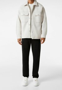 Vit ullblandad knäppjacka med två bröstfickor, bärs över en ljus topp och matchas med svarta cargo-byxor och vita sneakers.