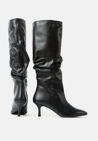 O pereche de cizme din piele negre, până la mijlocul gambei, cu vârful ascuțit, cu trunchiuri căzute, fermoare la spate și tocuri subțiri de tip kitten, pe un fundal alb simplu.