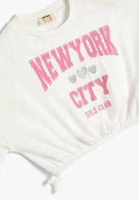 Camiseta corta blanca con dobladillo elástico, que presenta un texto en negrita de color rosa y plateado brillante que dice "CLUB DE CHICAS DE NUEVA YORK" con acentos de corazón.