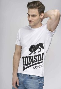 Balta medvilnės marškinėliai su dideliu juodu liūto grafiku ir ryškiu "Lonsdale London" tekstu. Trumpi rankoviai, apvalus iškirpimas, standartinė forma.