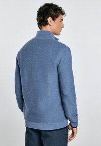 Uomo che indossa un maglione blu con un colletto alto, visto da dietro. Lo sfondo è grigio chiaro, mettendo in risalto i dettagli del capo.