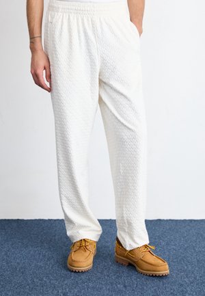 Personne portant un pantalon blanc ample texturé et des bottines beige à lacets, debout sur un tapis bleu les mains dans les poches.